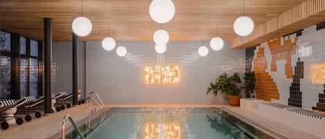 Una piscina interior moderna con techo de madera, paredes con azulejos y sillones de descanso.