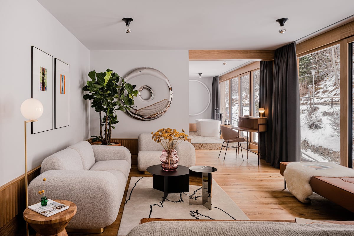 Ein modernes Wohnzimmer mit einer großen Pflanze, einem runden Spiegel und Blick auf eine verschneite Landschaft durch große Fenster.