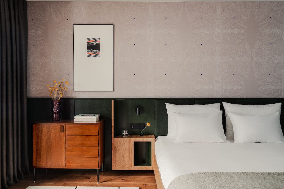 Ein modernes Hotelzimmer mit einem Holzkoffer, einem Nachttisch mit einer Vase, einem gerahmten Bild an der Wand und einem ordentlich bezogenen Bett.