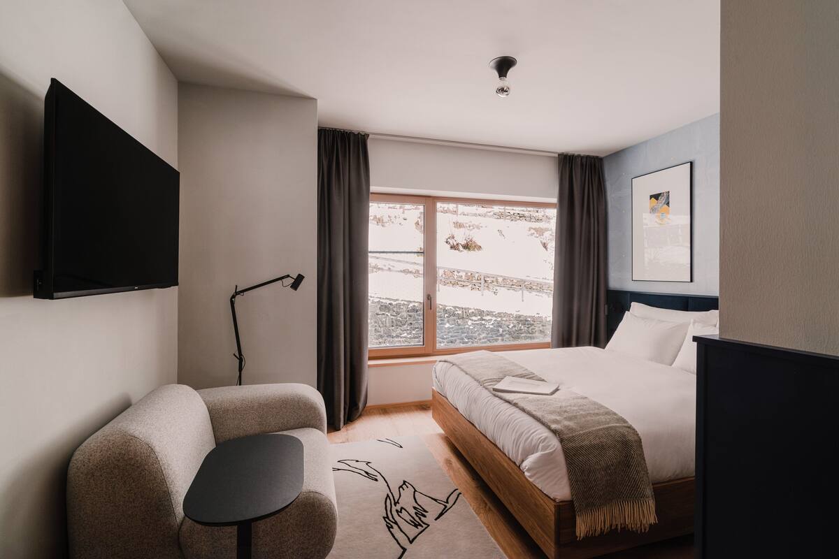 Ein modernes Hotelzimmer mit Bett, Schreibtisch, Sessel, Flachbildfernseher, einem Fenster mit Vorhängen und Blick auf die Stadt.