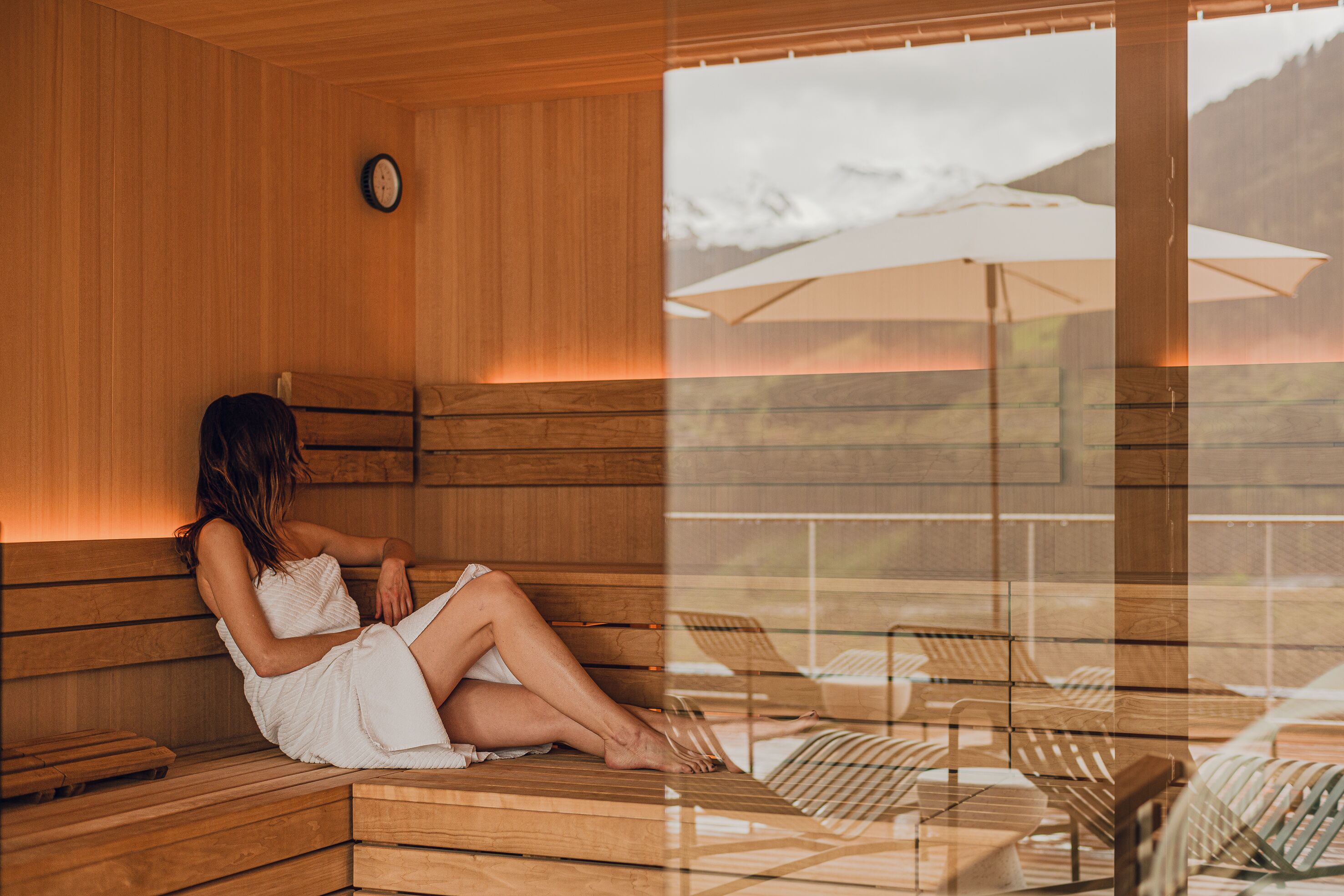 Un sauna en bois avec un banc, une horloge et une vue sur un espace extérieur aménagé avec des sièges et sur les montagnes.