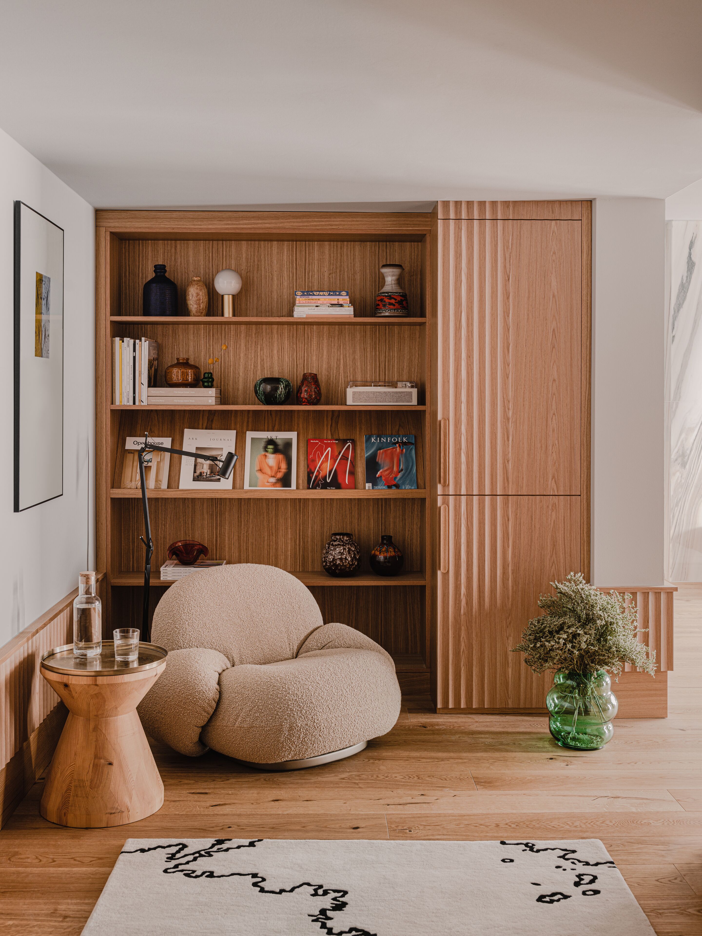 Una habitación acogedora con una estantería de madera, un sillón cómodo, una mesita auxiliar y un jarrón decorativo.