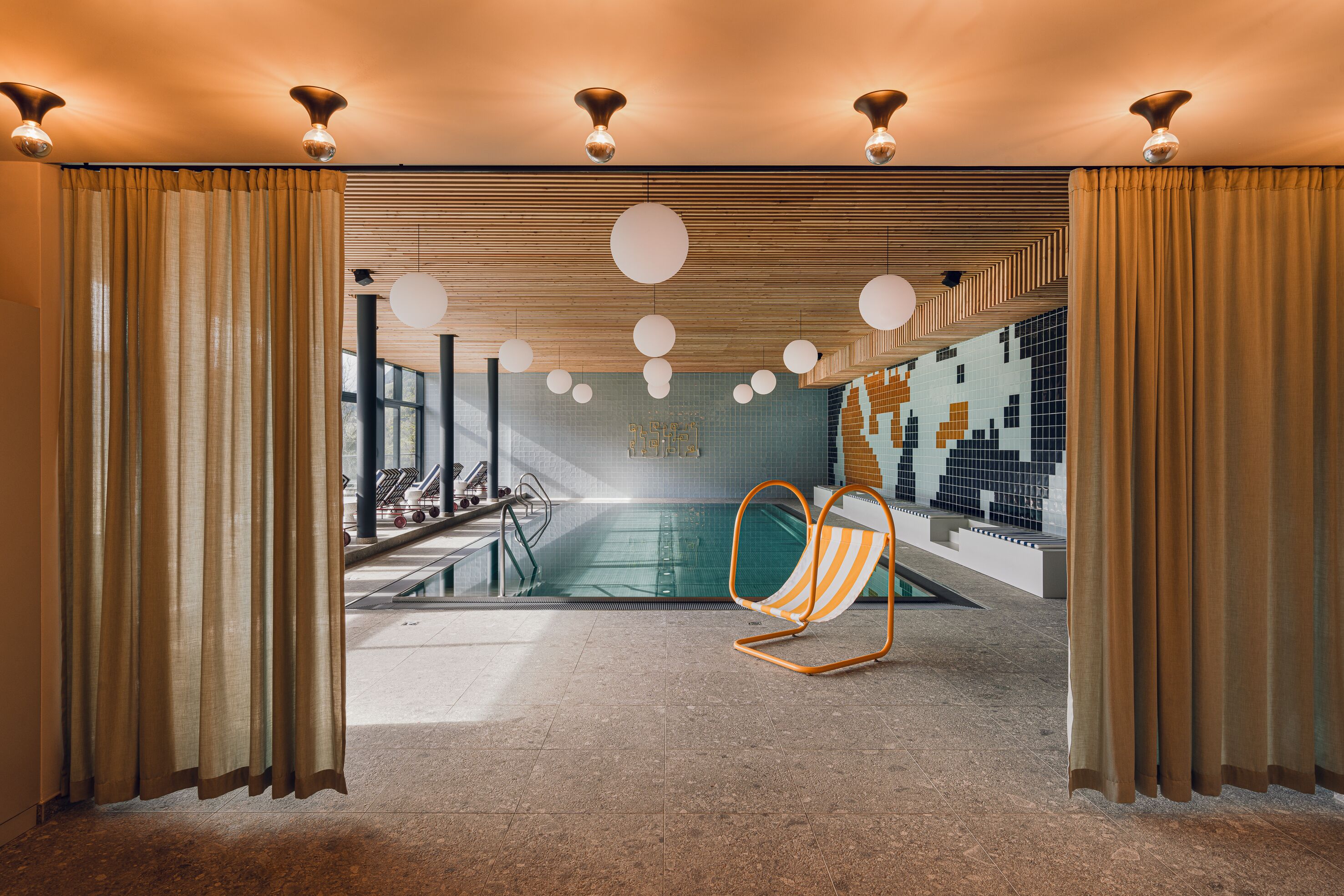 A modern indoor pool area with a clear view of the pool, a lounge chair, and a patterned wall.