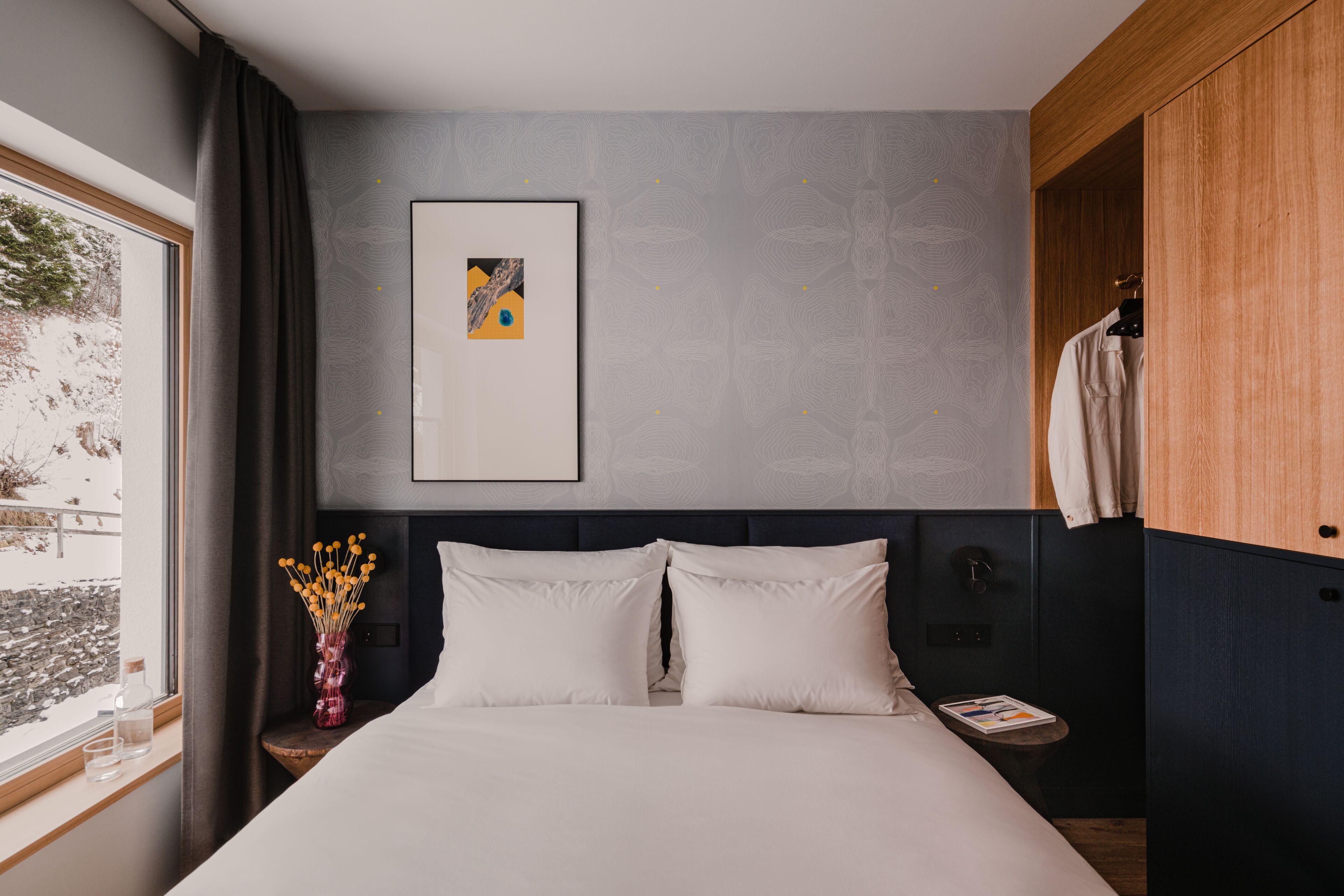 A modern hotel room with a large bed, a bedside table with a vase of flowers, a framed picture on the wall, and a window with curtains.