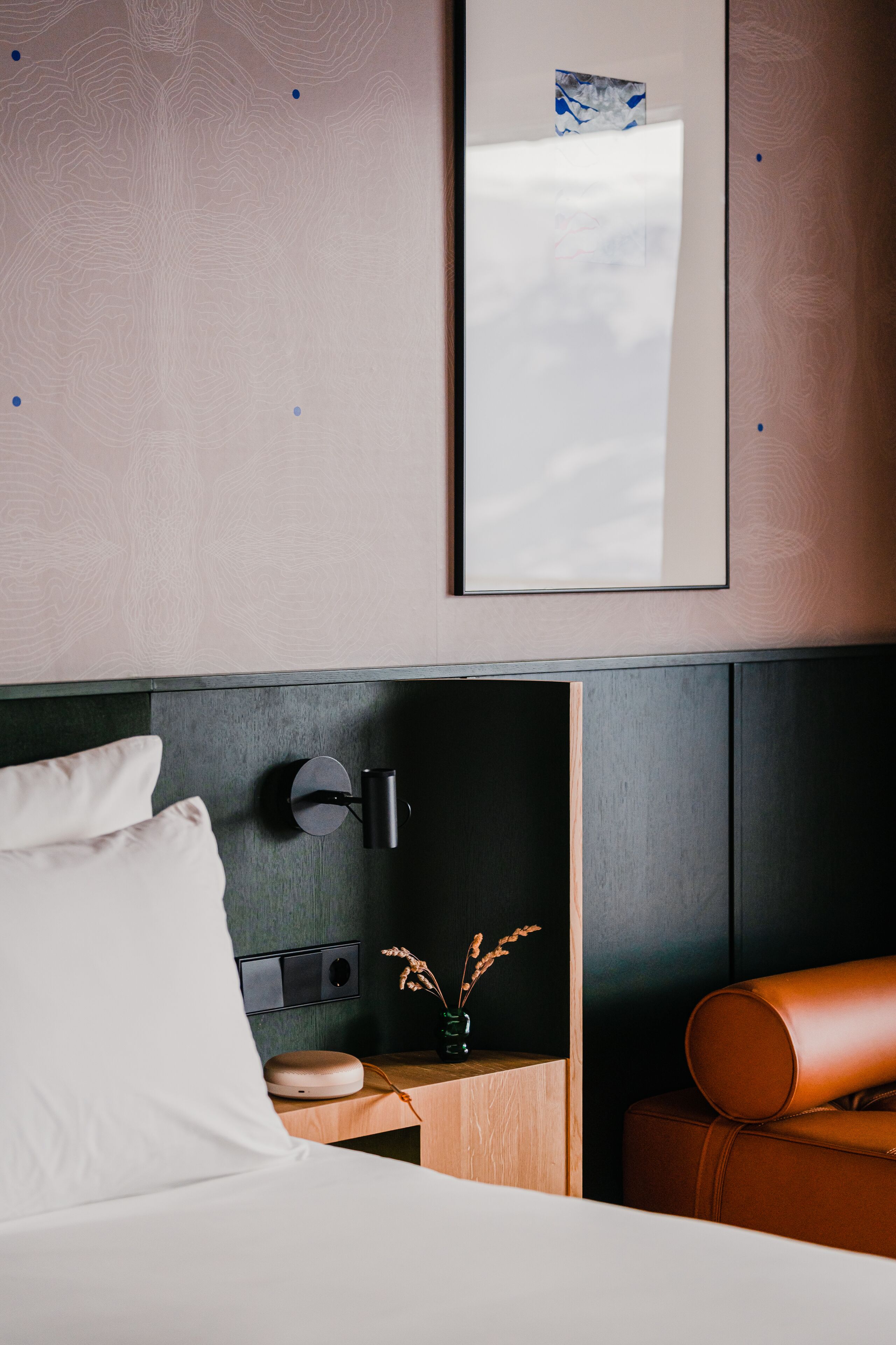 Ein modernes Hotelzimmer mit einem Bett, einem Nachttisch mit Lampe, einem Spiegel und einem Ledersessel.