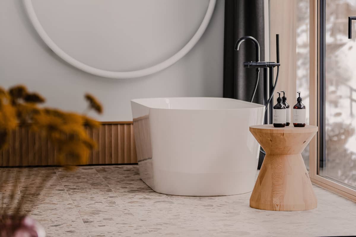 Ein modernes Badezimmer mit einer freistehenden Badewanne, einem hölzernen Beistelltisch mit Flaschen und einem großen Fenster.