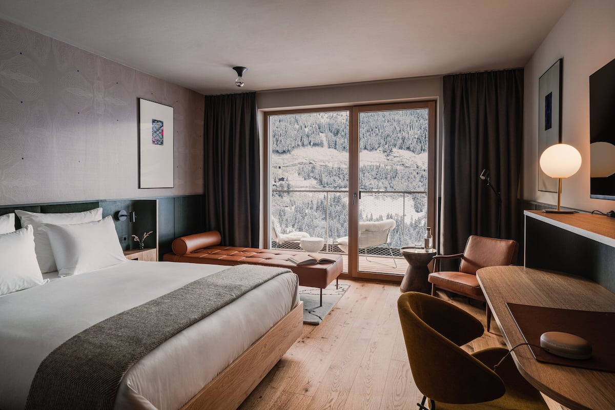 Ein Hotelzimmer mit Bett, Schreibtisch, Stuhl, einem Fenster mit Blick auf einen verschneiten Berg und einer Deckenleuchte.