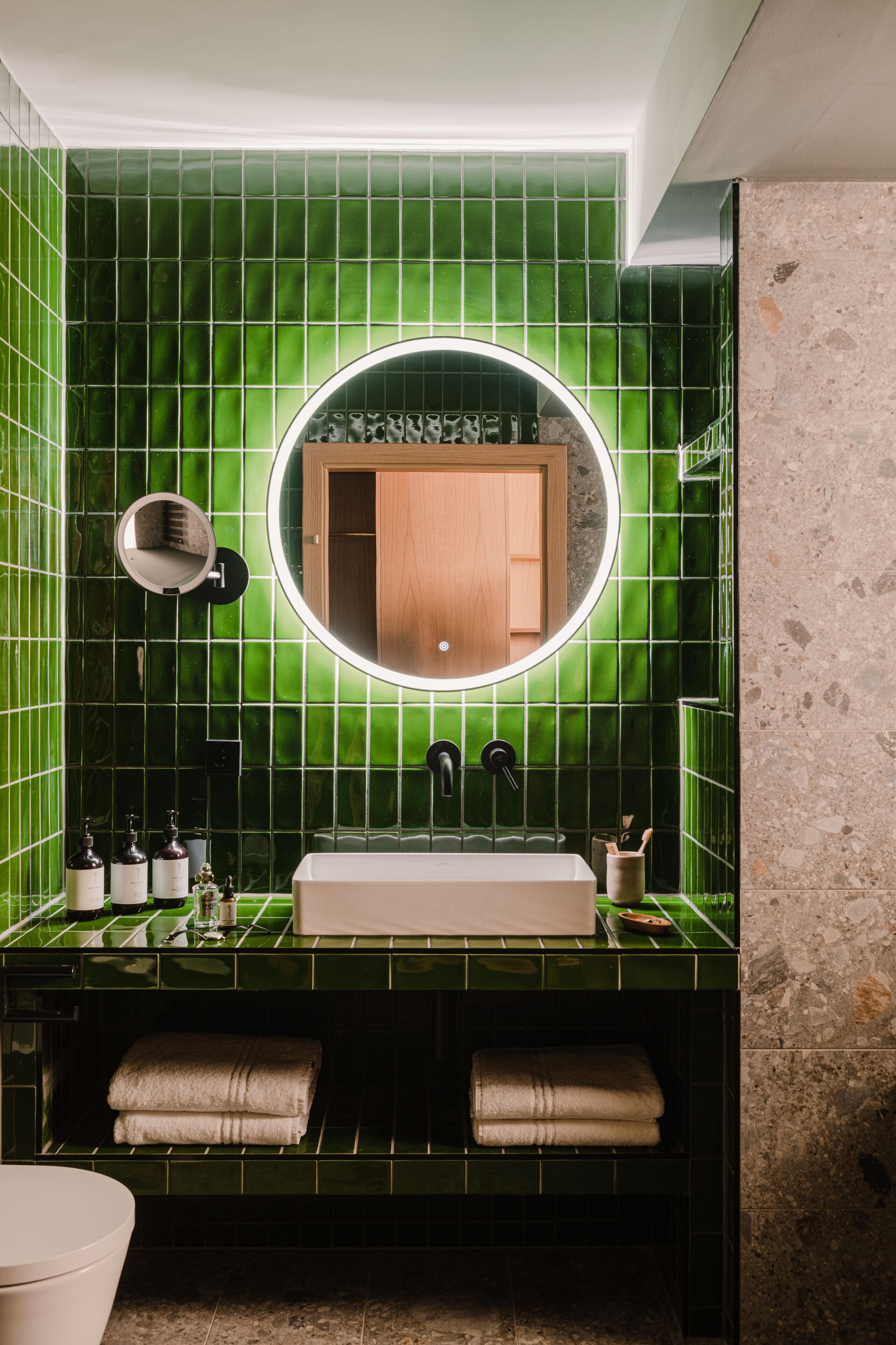 A bathroom with a green tiled wall, a round mirror, a wooden cabinet, a sink, and two towels.