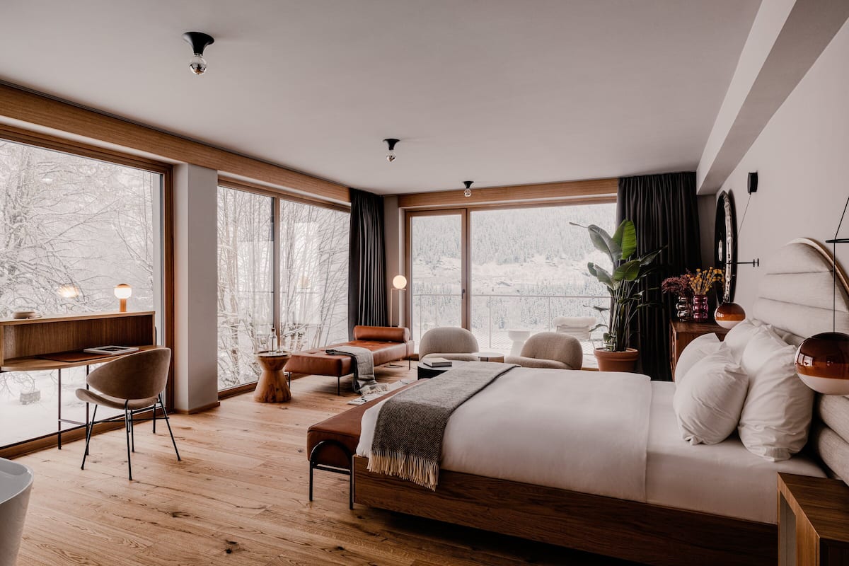 Ein modernes Schlafzimmer mit einem großen Bett, einem Schreibtisch und einem Stuhl. Große Fenster bieten Blick auf Bäume und eine verschneite Landschaft.