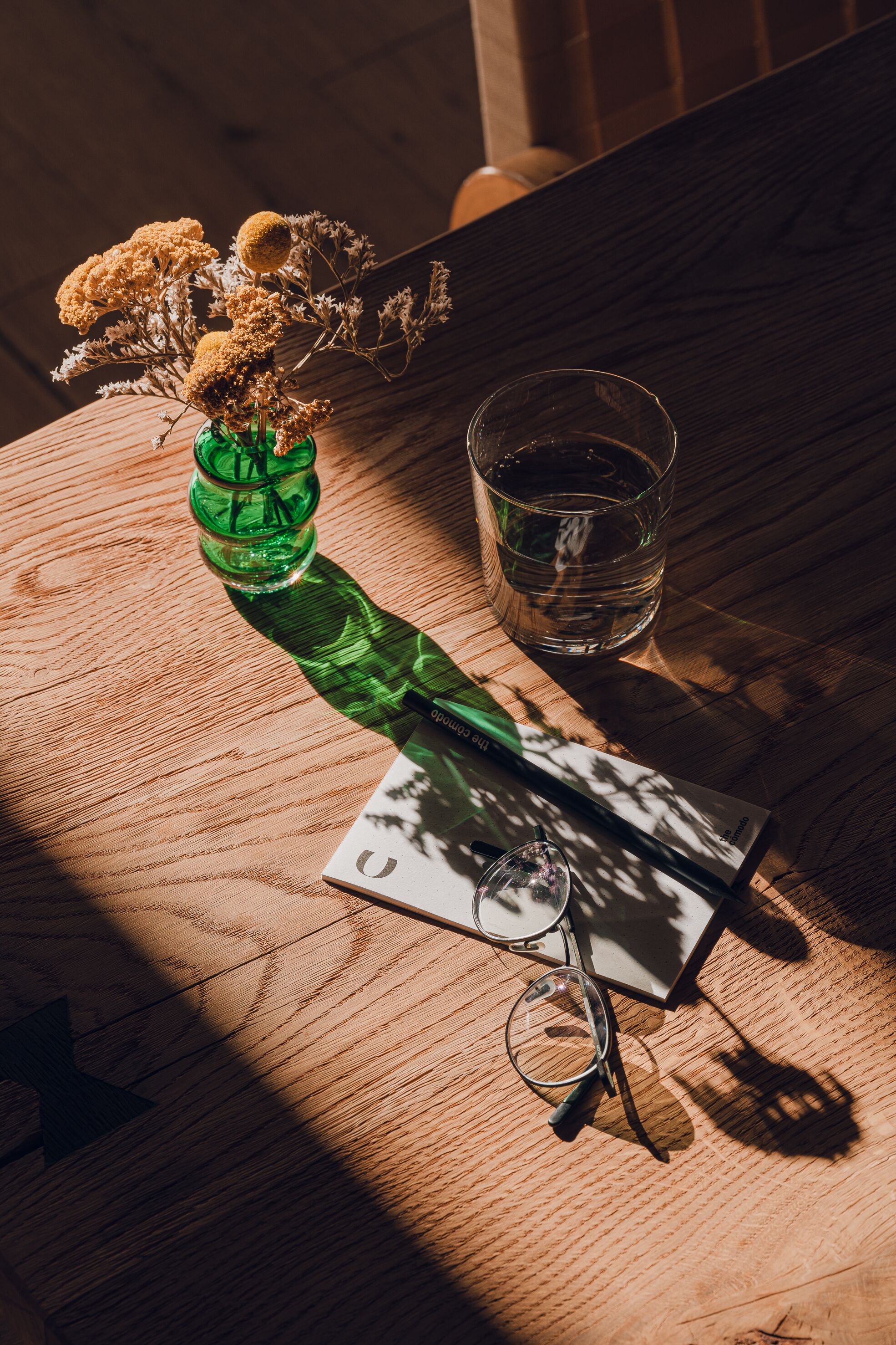 Ein Holztisch mit einer grünen Flasche, einem Glas Wasser, einem Brillenpaar und getrockneten Blumen.