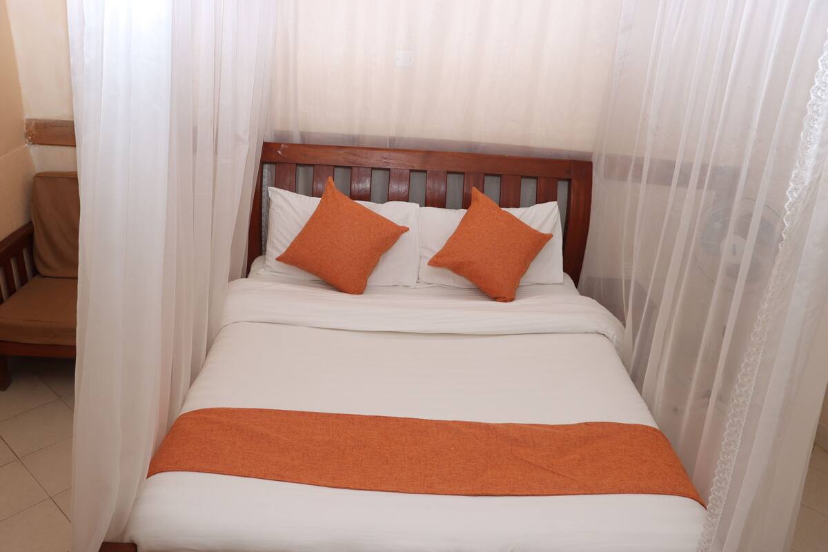 Cama com roupa de cama branca e duas almofadas laranja, cabeceira de madeira e colcha branca.