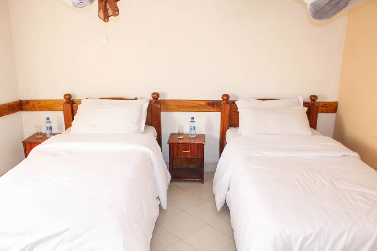 Quarto de hotel com duas camas de solteiro, cabeceiras de madeira, roupa de cama branca e mesas de cabeceira com garrafas de água.