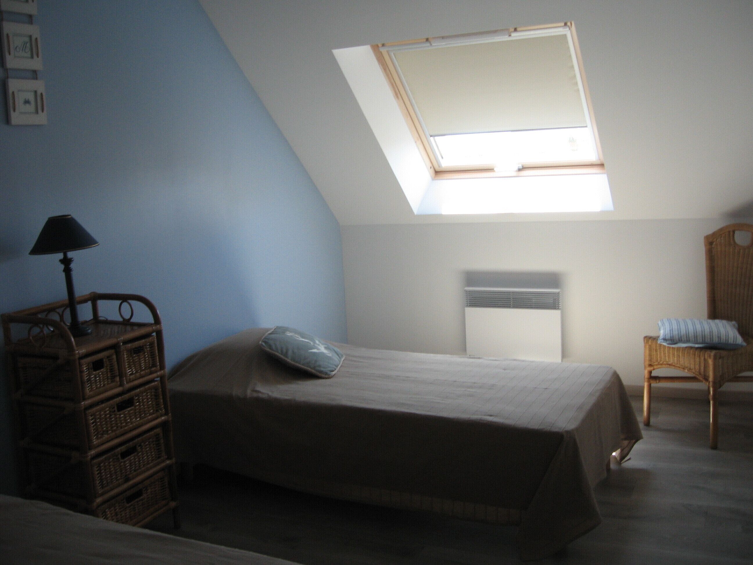 A bedroom with a sloped ceiling, a skylight, a bed, a chair, and a small table with a lamp.