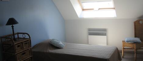 A bedroom with a sloped ceiling, a skylight, a bed, a chair, and a small table with a lamp.