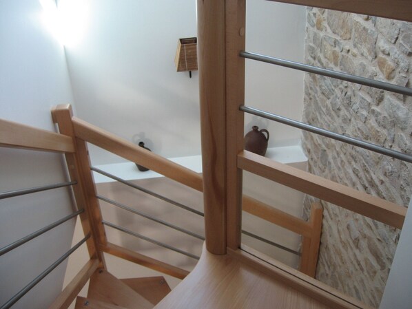 Un escalier en bois avec des rampes en métal mène à une salle de bain dont un des murs est en pierre et qui comprend une petite étagère sur laquelle est posé un pot en céramique.