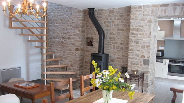 A dining area with a wooden table, chairs, a vase of flowers, a fireplace, and a kitchen in the background. - Gite Andrée, 800m from the beach in Pirou, garden, terrace, wifi, wood stove (Pirou)