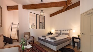 Ein Schlafzimmer mit einem Bett, zwei Nachttischen, einem Sessel, einem kleinen Tisch mit einer Vase, einer Pendelleuchte und einem gemusterten Teppich.