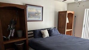 A bedroom with a bed, a wooden wardrobe, a shelf with a vacuum cleaner, and a framed picture on the wall. - Sun Lakes Country Club (Sun Lakes)
