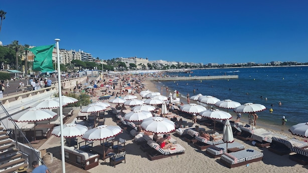 Een strand met veel ligbedden en parasols, een boulevard met mensen en een helderblauwe lucht.
