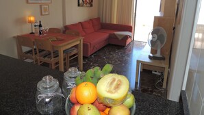 Un ambiente compatto con zona pranzo, un divano rosso, un tavolino con lampada e una ciotola di frutta sul bancone.