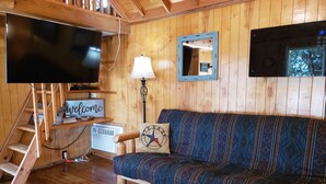 A wooden cabin interior with a sofa, a TV, a lamp, a welcome sign, and a staircase. - The Aoudad Cabin at Palo Duro  (Canyon)