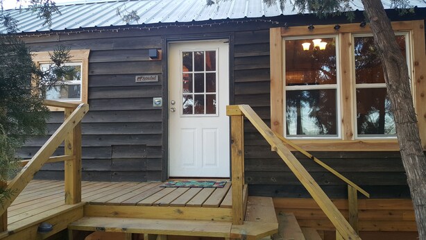 A wooden cabin with a white door, a small porch, and a window with a view of trees. - The Aoudad Cabin at Palo Duro  (Canyon)