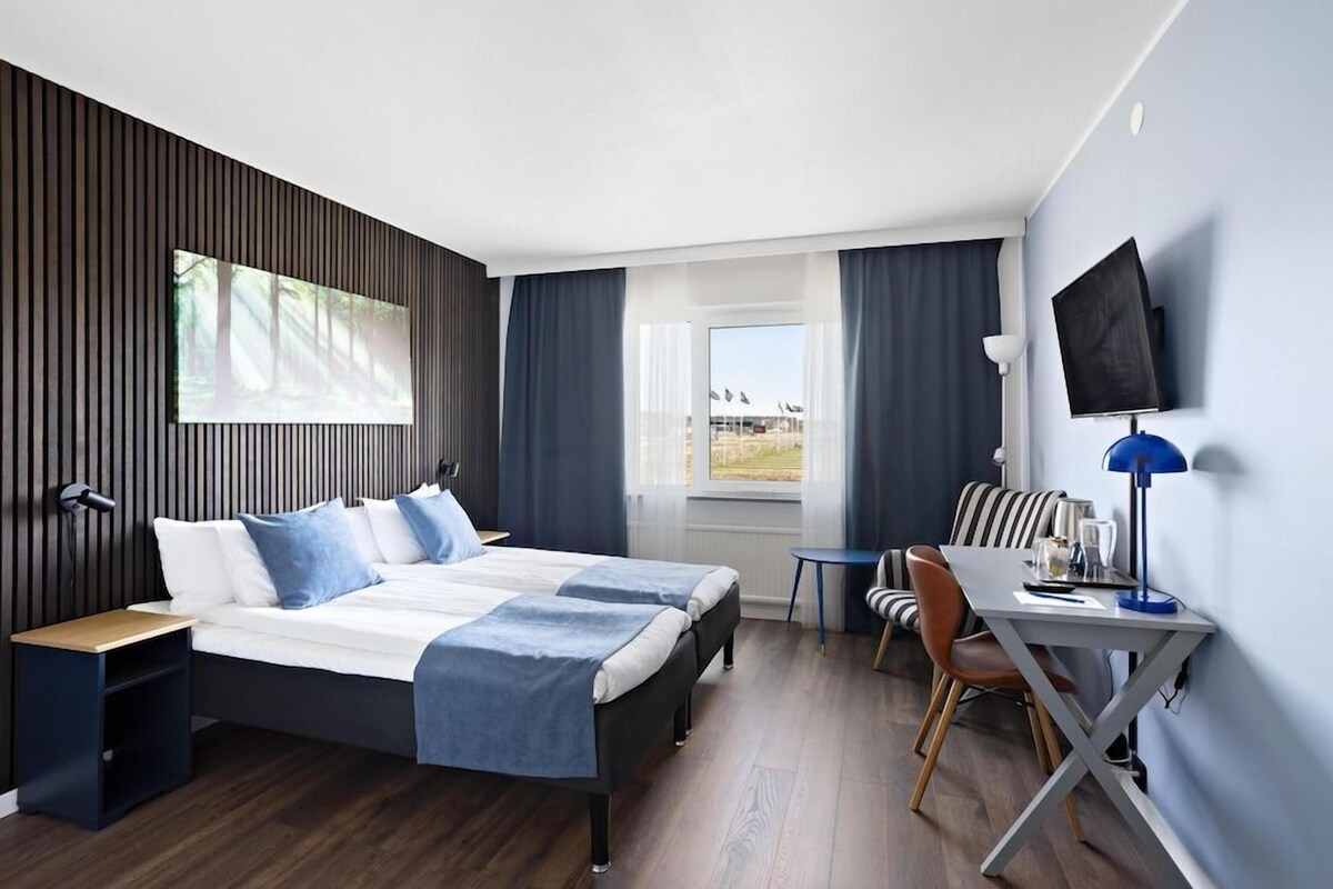 Una habitación de hotel moderna con una cama grande, un escritorio, una silla, un televisor y una ventana con vistas.