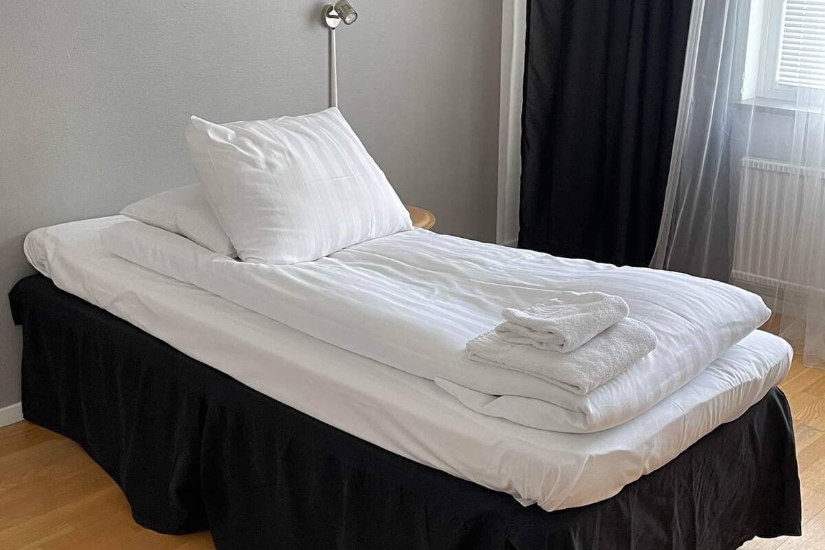 A neatly made bed with white linens, a black skirt, and a bedside lamp.