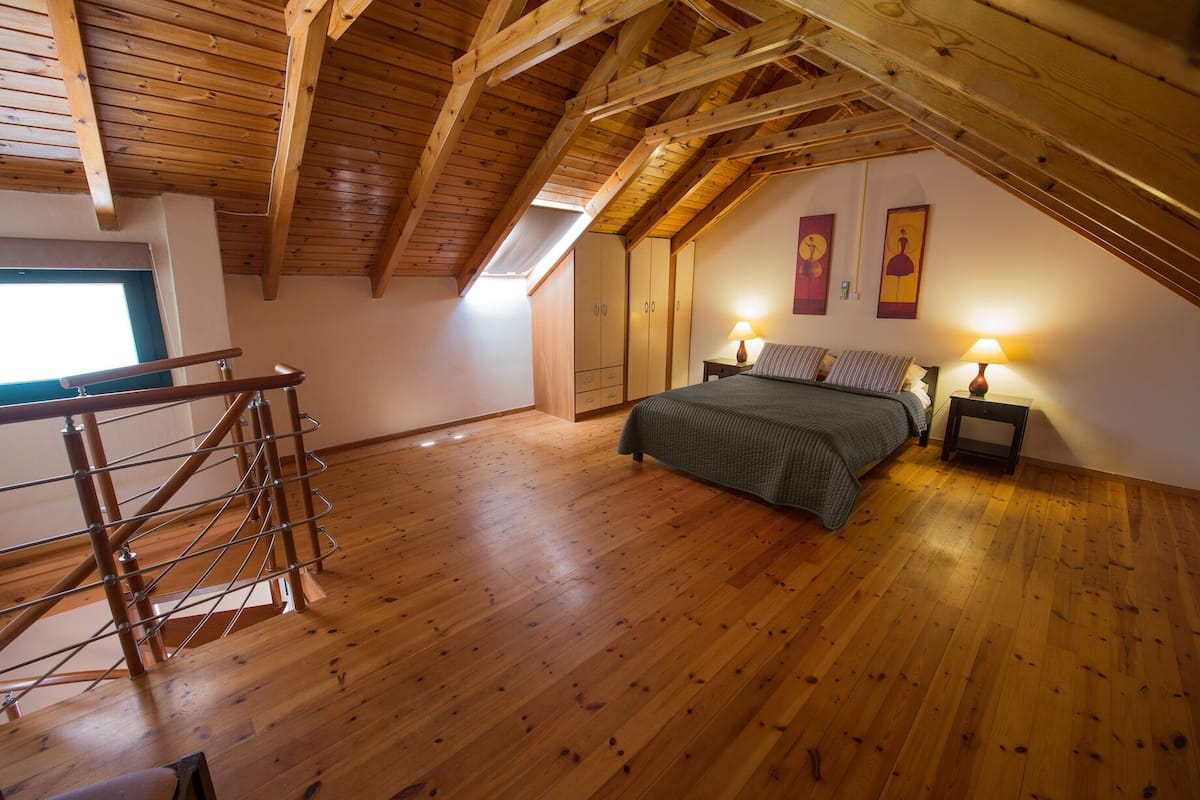 A bedroom with a sloped ceiling, wooden flooring, a bed with pillows, two bedside lamps, and a window.