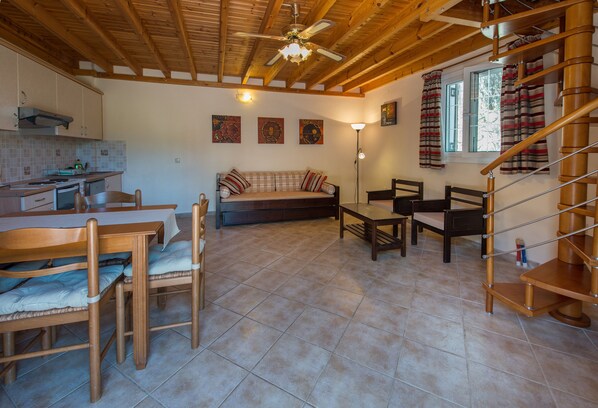 A cozy kitchen with wooden cabinets, a dining table, and chairs. There is a sofa with cushions and a floor lamp. A staircase with a wooden railing is visible.