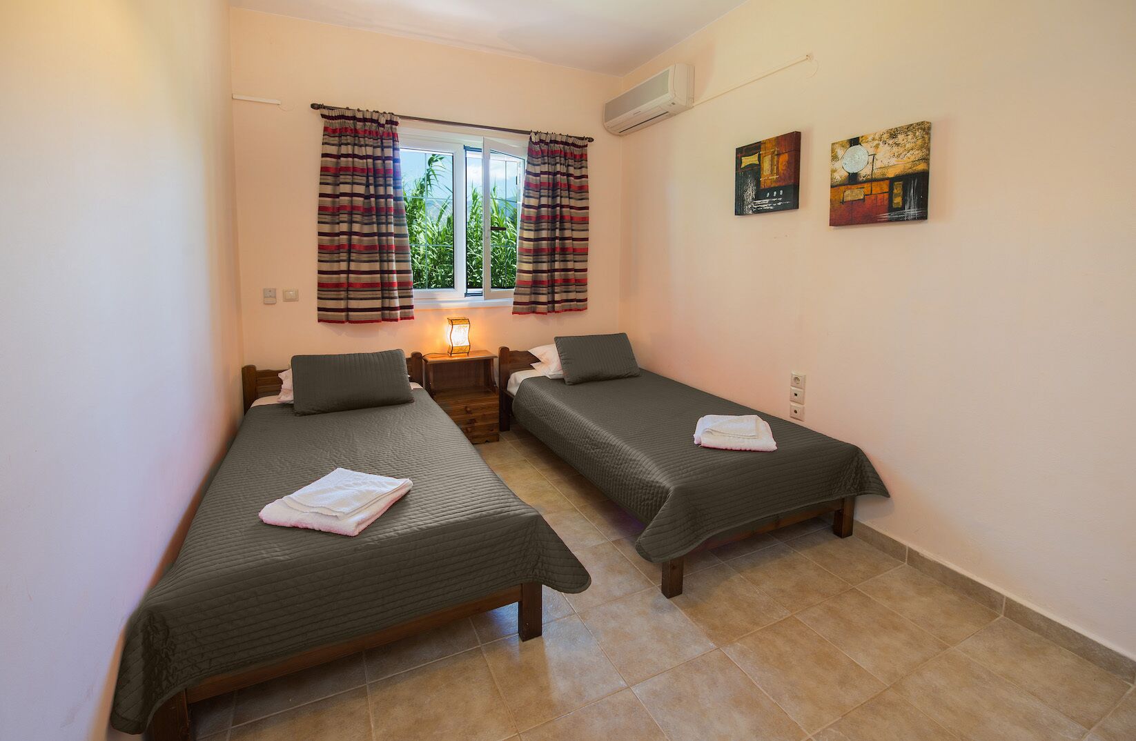 A room with two single beds, a nightstand with a lamp, a window with curtains, and two paintings on the wall.