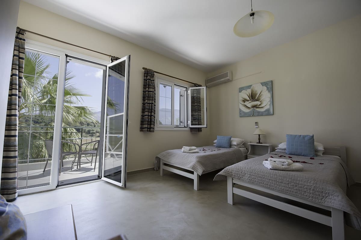 A bedroom with two beds, a balcony with a view of palm trees, and a painting on the wall.