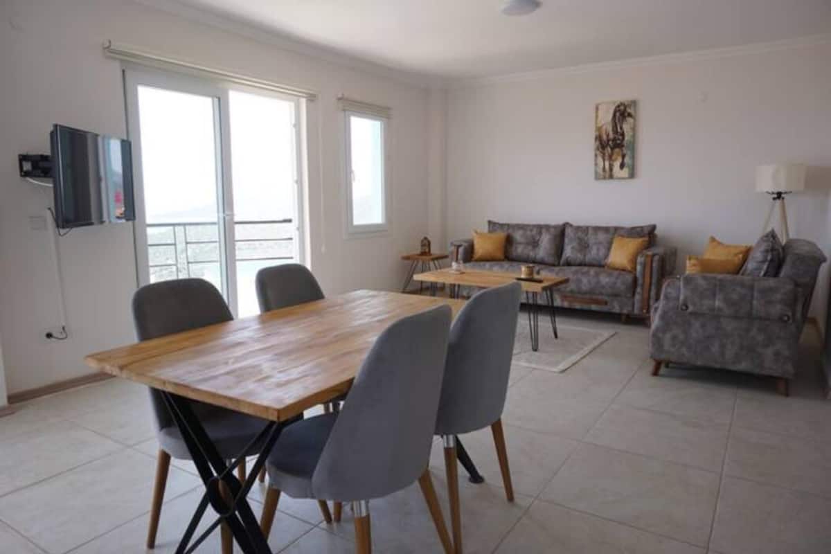 Amplia sala de estar con una mesa de comedor de madera, sillas grises, un sofá gris con cojines amarillos y un televisor montado en la pared.