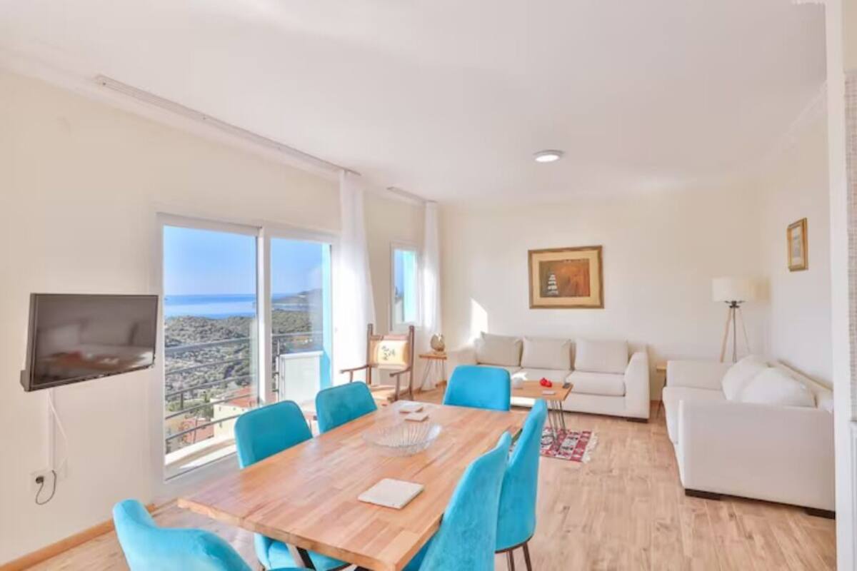 Amplia sala de estar con un ventanal grande, un televisor de pantalla plana montado en la pared, una zona de comedor con una mesa de madera y sillas azules, y un sofá blanco.
