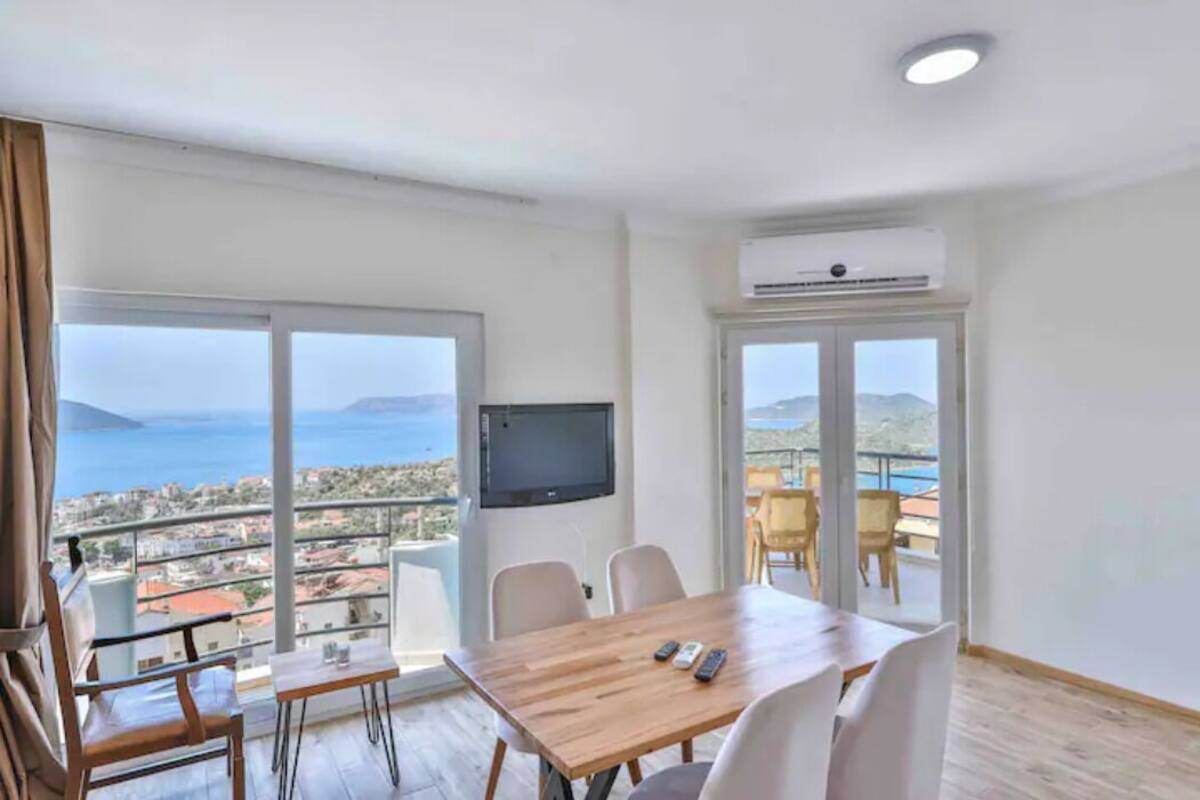 Una habitación con una mesa de madera, sillas, un televisor y ventanas amplias con vistas al mar y a la ciudad.