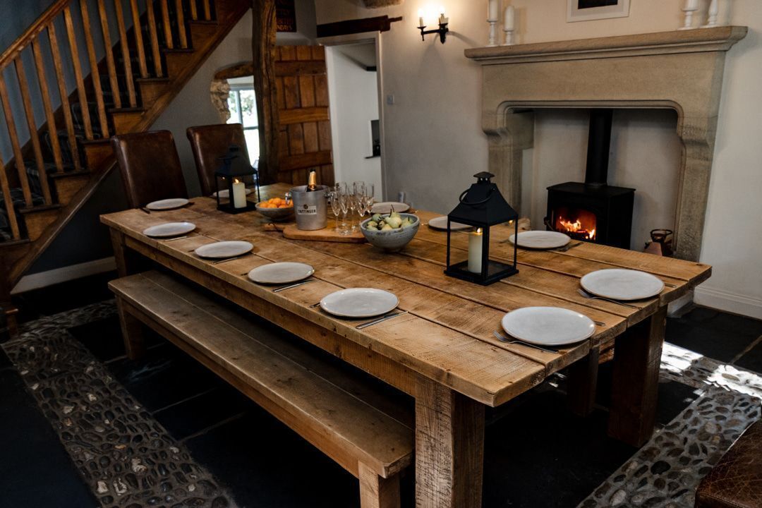 A wooden dining table set with plates, glasses, and a bowl of fruit, with a fireplace and staircase in the background.