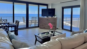 A spacious living room with a large sofa, a glass coffee table, and a television. The room has large windows offering a view of the beach and ocean. - Beach Front,2bed 2bath, Pool, Huge Balcony (panama city beach)