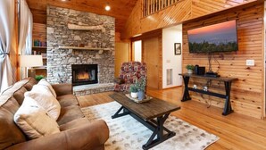 A cozy living room with a stone fireplace, a comfortable sofa, a coffee table, and a television mounted on the wall. - Incredible Mountain Views Near Dtown Gatlinburg! (Gatlinburg)