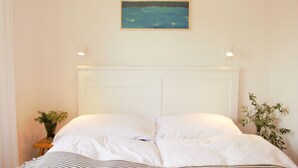 Un dormitorio con cabecera blanca, cama con ropa de cama blanca, un cuadro de una escena marina y dos mesitas de noche con lámparas y plantas.