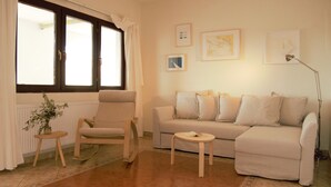 Una sala de estar con un sofá color beige, un sillón mecedor de madera, una mesita y una ventana con cortinas.