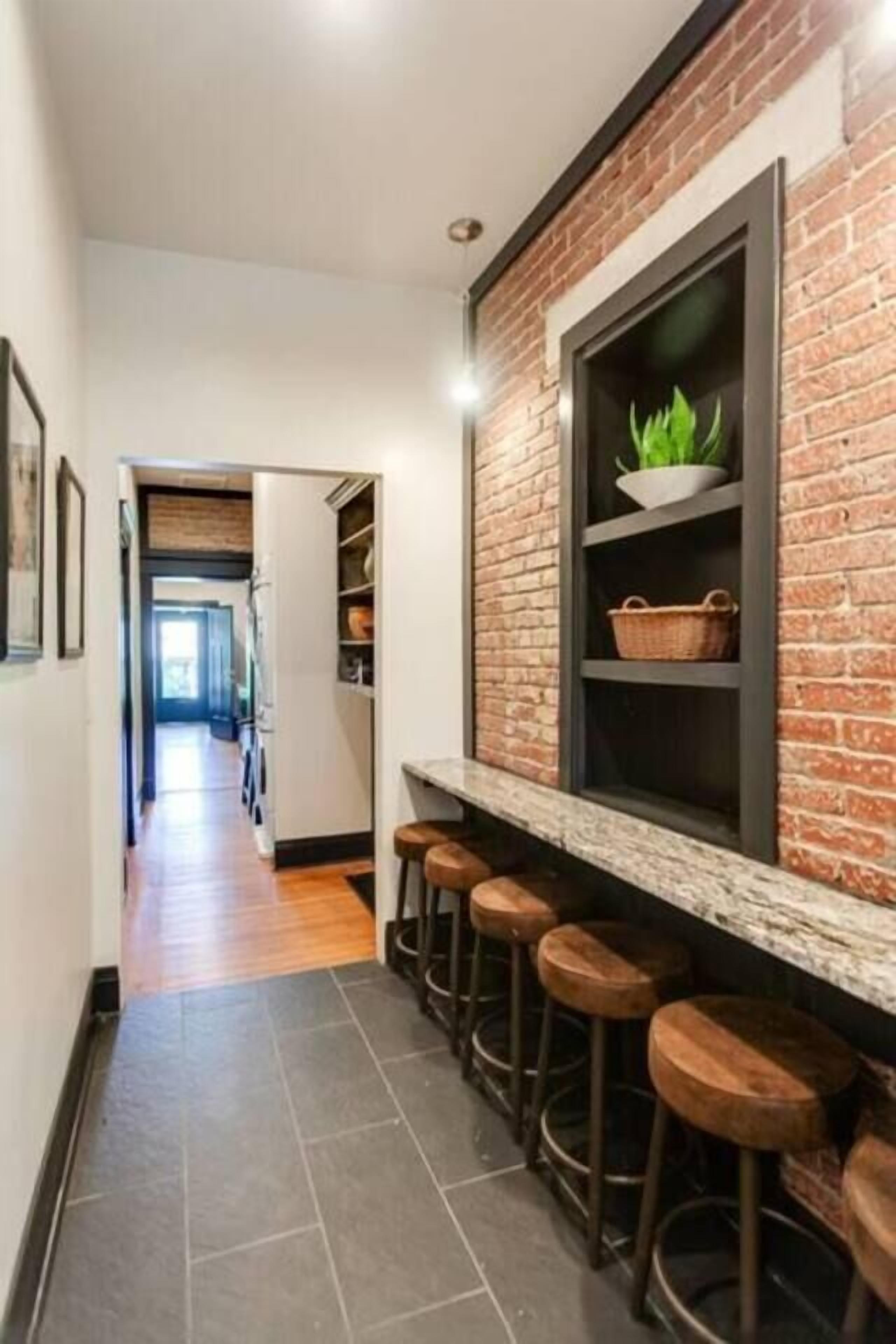 Un couloir étroit avec un mur en briques, une fenêtre ornée d’une plante et une rangée de tabourets en bois.