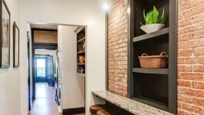 Un pasillo estrecho con una pared de ladrillo, una ventana con planta y una hilera de taburetes de madera.