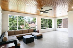 Relax in this serene space with a high ceiling, ceiling fan, and large windows offering views of the surrounding greenery.