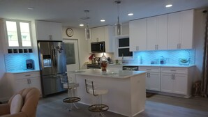 A modern kitchen with white cabinets, a stainless steel refrigerator, a microwave, and a sink. There are two bar stools and a clock on the wall. - Relax by the sea! Three bedroom home with pool awaits you near Destin Beaches. (Fort Walton Beach)