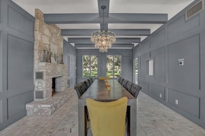 A spacious dining room with a long table and yellow chairs, a stone fireplace, and large windows with a view of greenery.