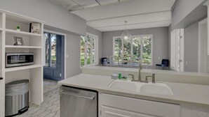 Cocina moderna con armarios blancos, lavavajillas de acero inoxidable, un lavamanos doble y una ventana con vista a zonas verdes.