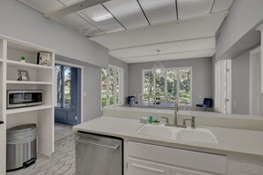 A modern kitchen with white cabinetry, a stainless steel dishwasher, a double sink, and a window with a view of greenery.