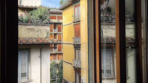 Vista pela janela de um edifício com várias varandas, algumas adornadas com plantas e flores.