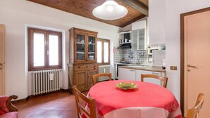Uma sala de jantar com uma mesa redonda coberta por uma toalha xadrez vermelha e branca, cadeiras de madeira, um armário com portas de vidro, uma área de cozinha com armários brancos e um revestimento de azulejos, e um teto de madeira.