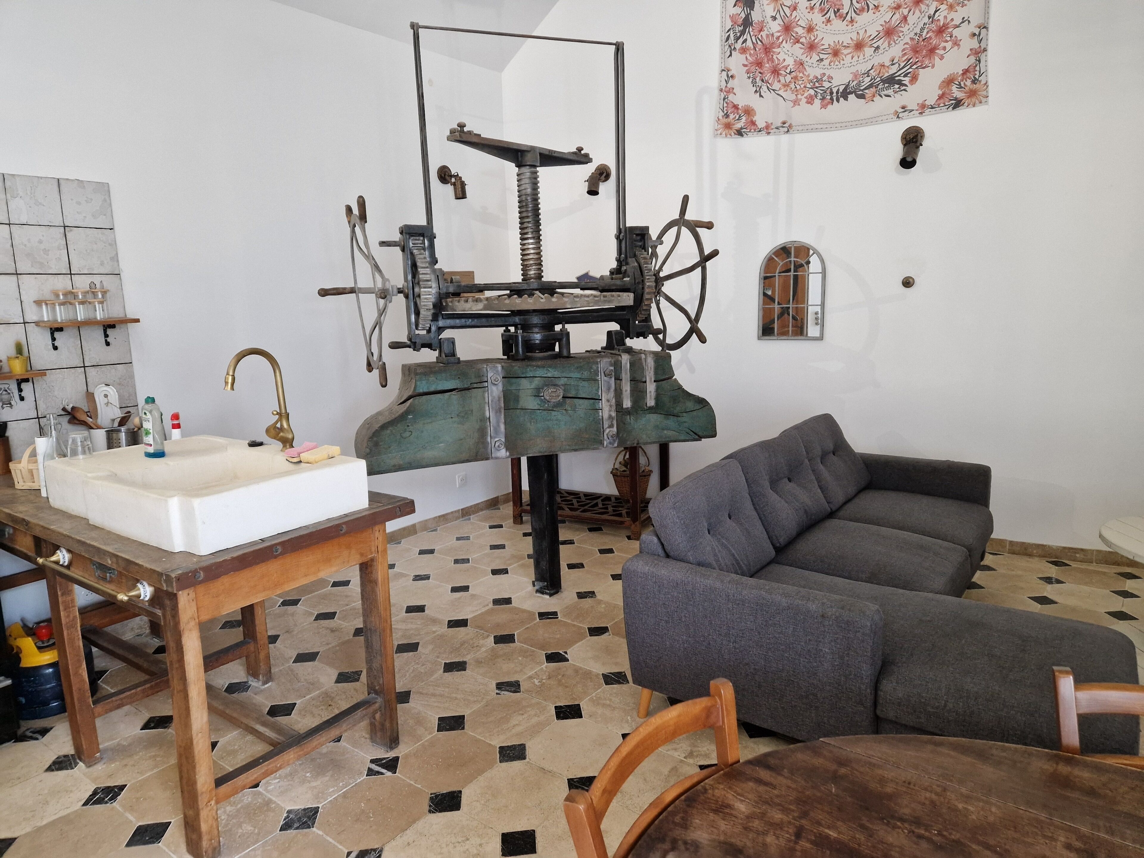 A room with a sink, a wooden table, a grey sofa, and a vintage sewing machine.
